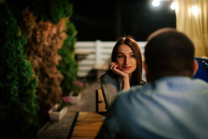 A woman with long brown hair smiling gently during a night conversation, discussing being conventionally attractive.