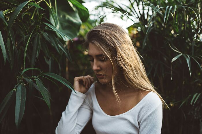 A woman with long blonde hair looking down amidst lush green foliage, reflecting on being conventionally attractive.