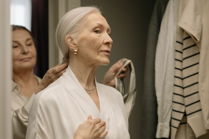 An older woman getting ready, another woman assisting her, exploring the realities of being conventionally attractive.