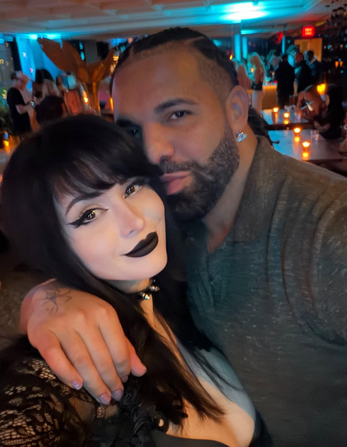 A Goth Baddie with dark hair and makeup poses with a bearded man, both looking at the camera, after a dating show. A Goth Baddie with dark hair and makeup poses with a bearded man, both looking at the camera, after a dating show.