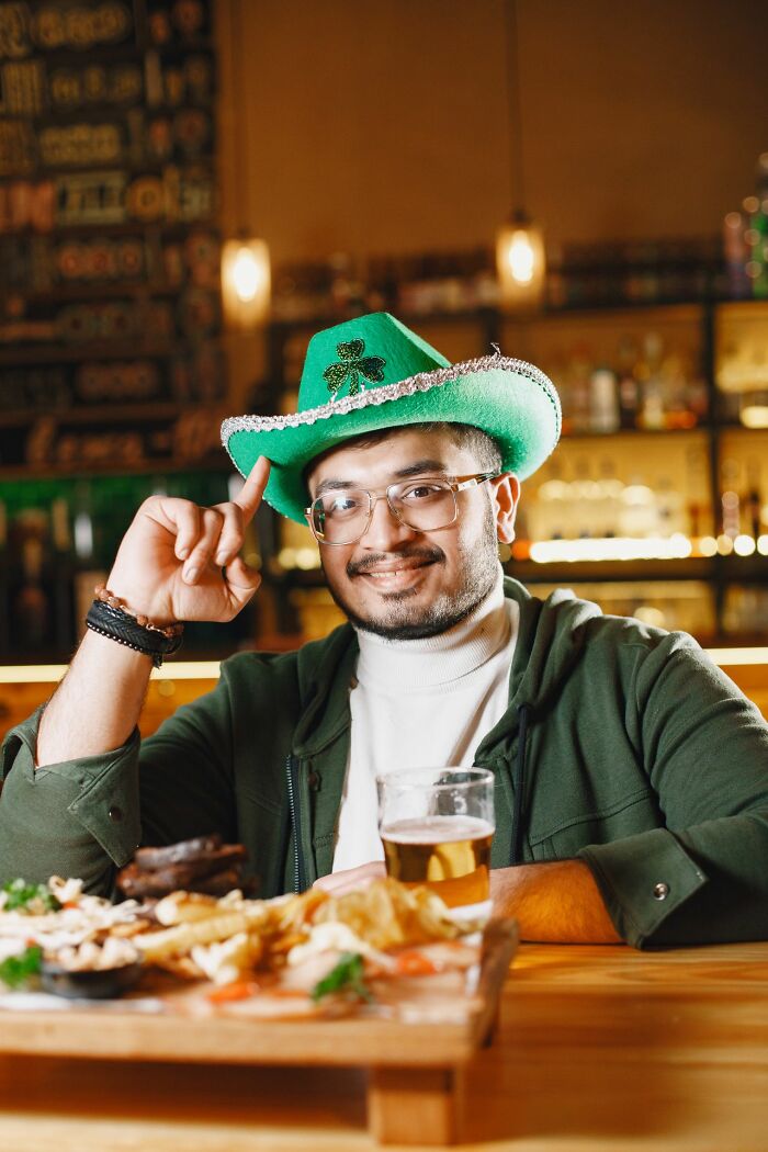 A man wearing a green hat and smiling, enjoying a meal and beer, a reminder for Americans to be respectful abroad.