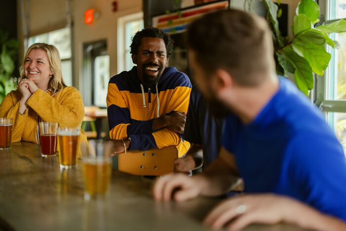 Three friends laughing at a bar with drinks, reminding Americans to be respectful when visiting other countries.