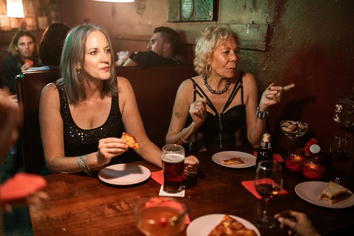 Two American women at a bar, eating pizza and smoking, a scene that reminds Americans to be respectful in other countries.