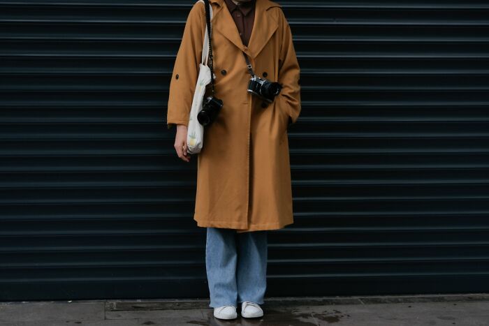 Person in a trench coat with cameras and a tote bag, representing pink taxes making being a woman more difficult.
