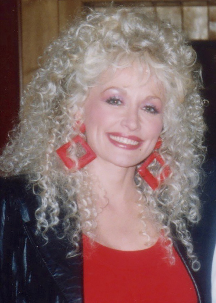 Young Dolly Parton smiling with big curly blonde hair and large red square earrings, writing a sweet letter to Eminem.