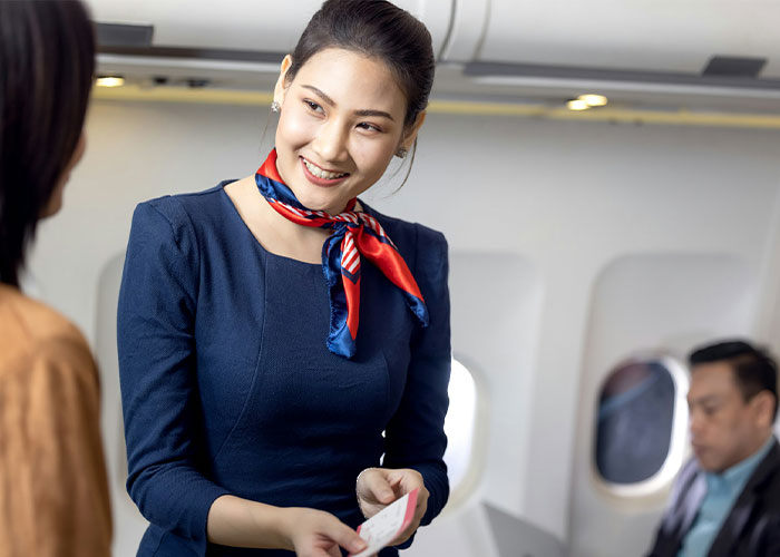 A smiling flight attendant in a blue uniform and red scarf speaks to a passenger about a plane seat issue. A smiling flight attendant in a blue uniform and red scarf speaks to a passenger about a plane seat issue.