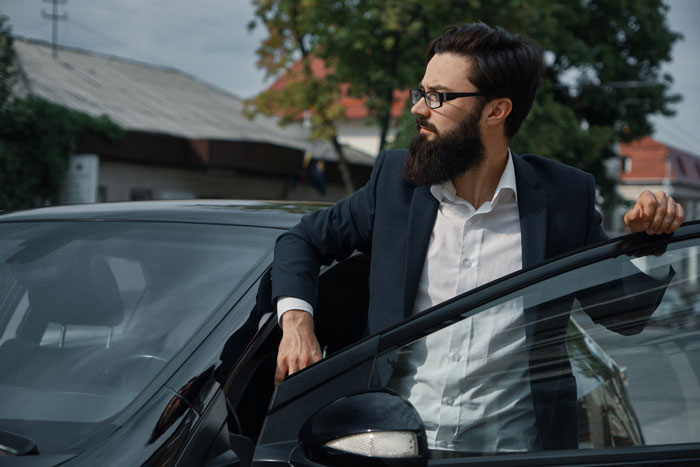 A man with a beard and glasses getting out of a black car, looking like he might throw a fit.