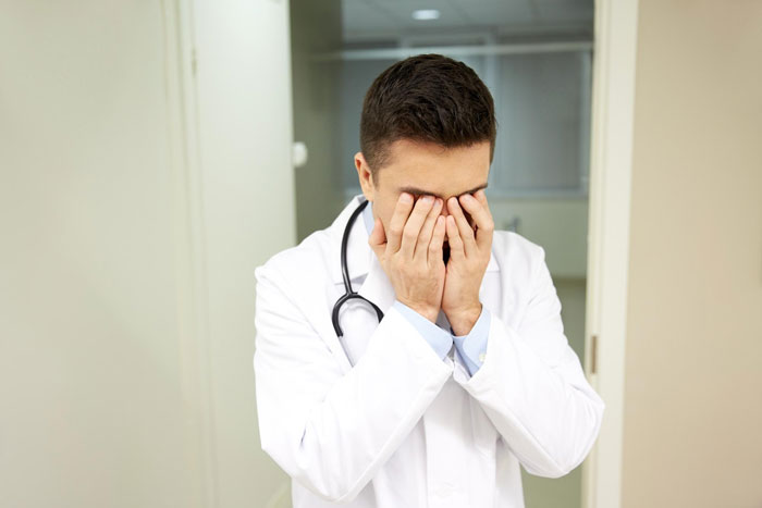 A medical worker in a lab coat and stethoscope, looking distressed and covering his face with his hands. A medical worker in a lab coat and stethoscope, looking distressed and covering his face with his hands.