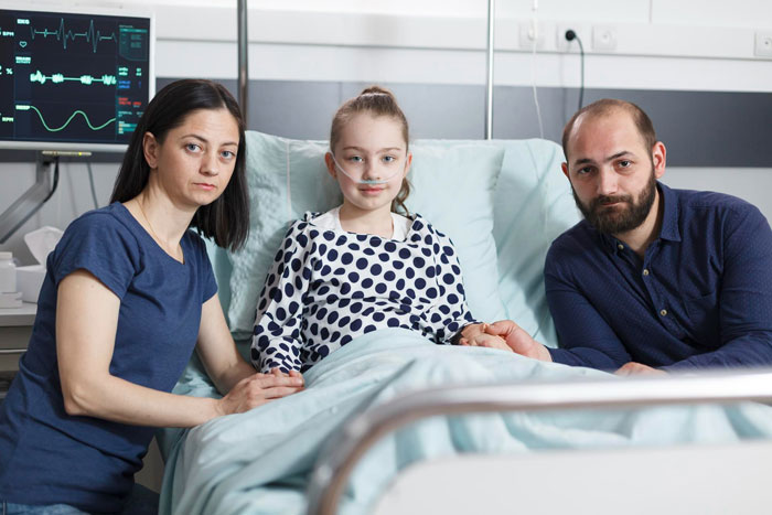 A mom and dad look concerned while their daughter, a patient, rests in a hospital bed with a medical worker in the background. A mom and dad look concerned while their daughter, a patient, rests in a hospital bed with a medical worker in the background.