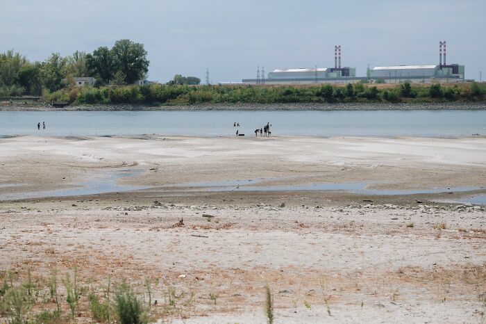 A riverbed suffering from a brutal drought in Europe, exposing dry land and shallow water with industrial buildings in background.