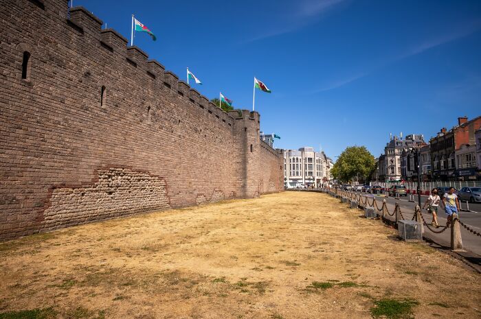 A historic castle wall in Europe overlooks parched brown grass, a visual impact of the brutal drought.
