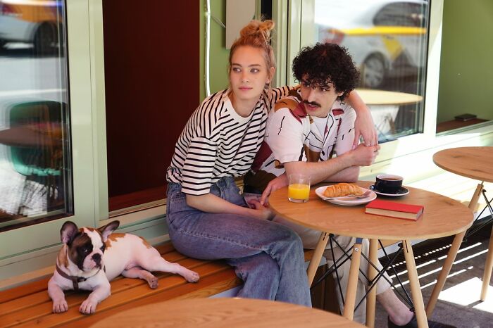 A couple sitting at an outdoor cafe with a dog, reflecting on funny questions Americans asked Europeans.