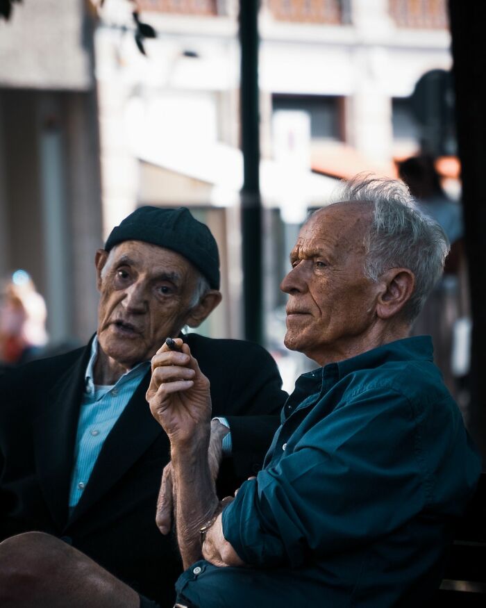 Two elderly men sitting on a bench, one with a beanie talking to the other. Americans asked Europeans many questions.