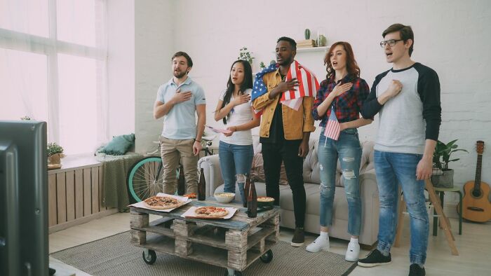 Diverse group of young adults standing in a living room, hands over hearts, watching a TV. Americans asked Europeans many questions.
