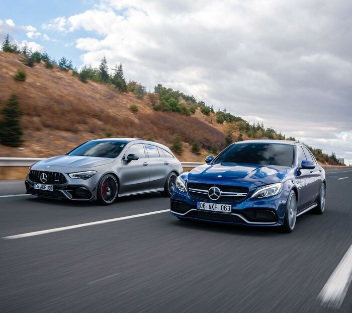 A grey station wagon and a blue sedan driving side-by-side on a highway with hills in the background. Americans asked Europeans questions about cars.