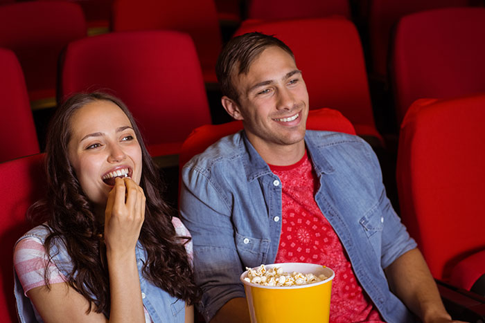 Happy couple, a guy and a girl, watching a movie in a theater. She is eating popcorn and smiling, while he is looking at her with a soft smile. They appear to be enjoying their time together before she changes her hair color.