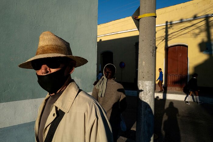 A man in a straw hat and mask walks down a street, a street photo from the Macadam Awards.