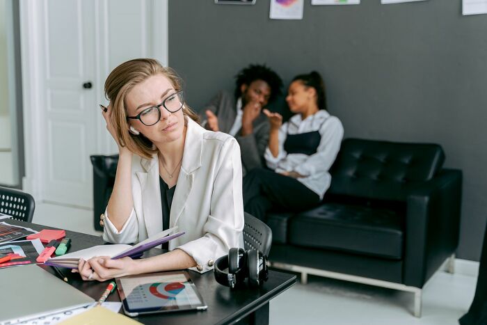 Woman at a desk looking upset while two people gossip behind her, questioning respect in their lives.
