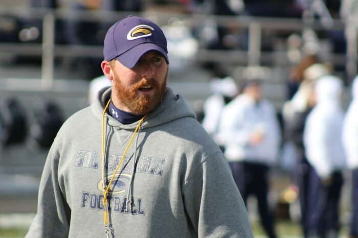A man with a beard, wearing a blue cap and a grey hoodie that says GOLDEN EAGLE FOOTBALL. People in their lives.