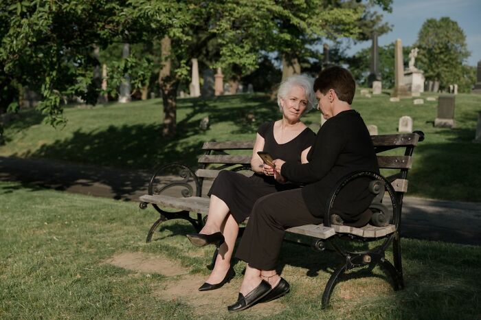 Two women sitting on a bench in a park-like setting, possibly a cemetery, looking at a phone. People in their lives.