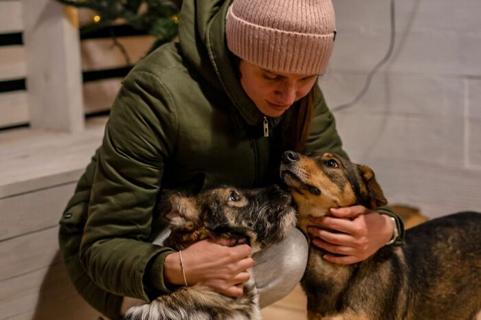 A woman wearing a pink beanie and a green jacket, lovingly embracing two dogs. People in their lives.