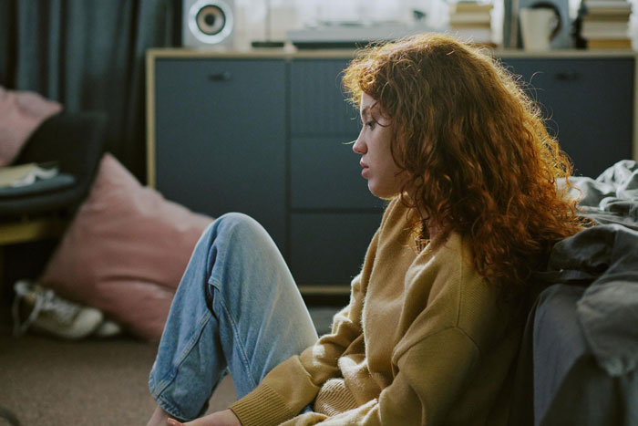 A woman with red, curly hair sitting on the floor, looking down, representing the father-daughter bond being ruined. A woman with red, curly hair sitting on the floor, looking down, representing the father-daughter bond being ruined.
