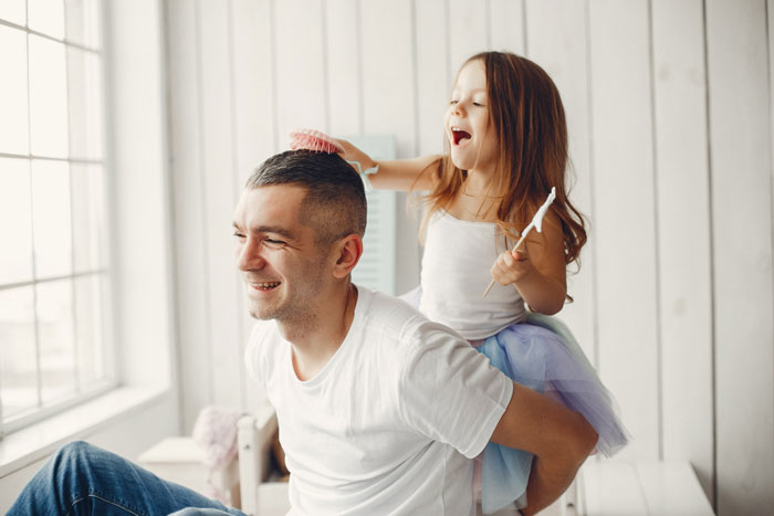 A happy young girl playfully brushes her dad's hair, symbolizing a normal father-daughter bond. A happy young girl playfully brushes her dad's hair, symbolizing a normal father-daughter bond.