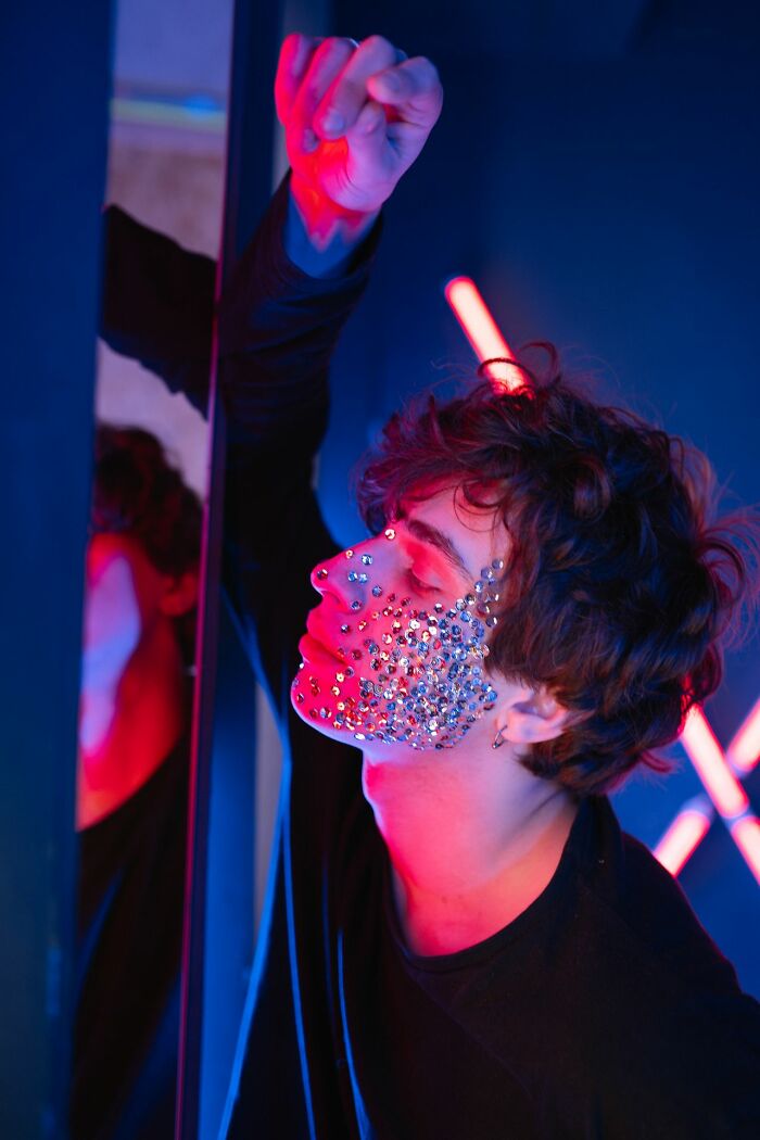 A person with sequins on their face, leaning against a mirror in a vibrant, neon-lit room, shutting down creepy men.