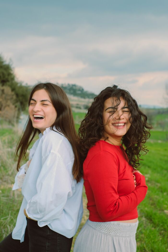 Two women laughing cheerfully, standing back-to-back in a green field, expressing joy and confidence.