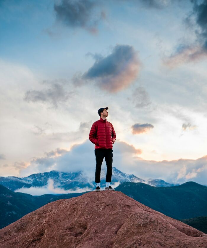 A man in a red jacket standing atop a hill, with mountains and a cloudy sky in the background, showing confidence.