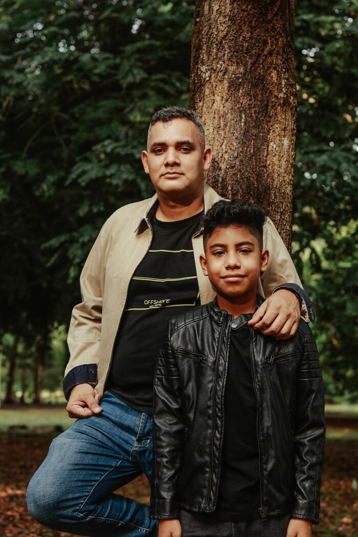 A father and son standing by a tree, looking at the camera, showing a bond.