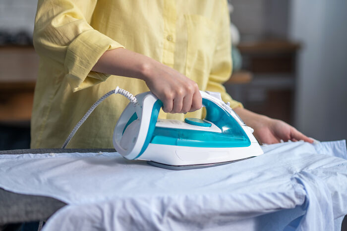 A person ironing a blue shirt on an ironing board with a white and blue iron. Ironing might be an unhinged house rule in a family.
