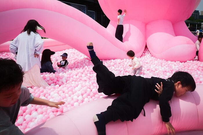 Children play in a large pink ball pit, with one child climbing on an inflatable structure, a street photo from the Macadam Awards.