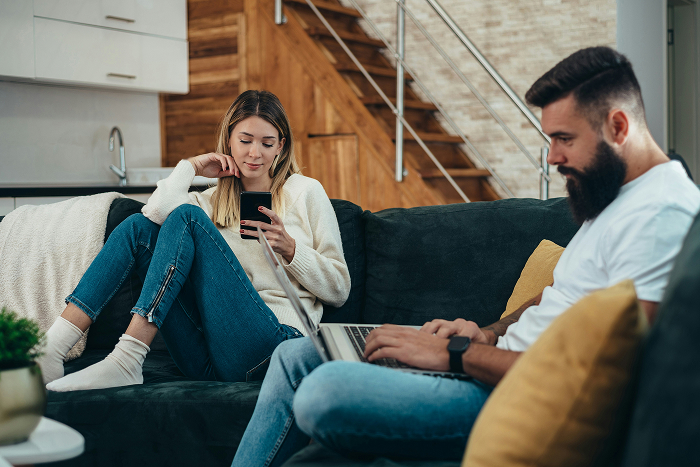 A couple sits on a couch, the man on a laptop and the woman on her phone, discussing an open relationship. A couple sits on a couch, the man on a laptop and the woman on her phone, discussing an open relationship.