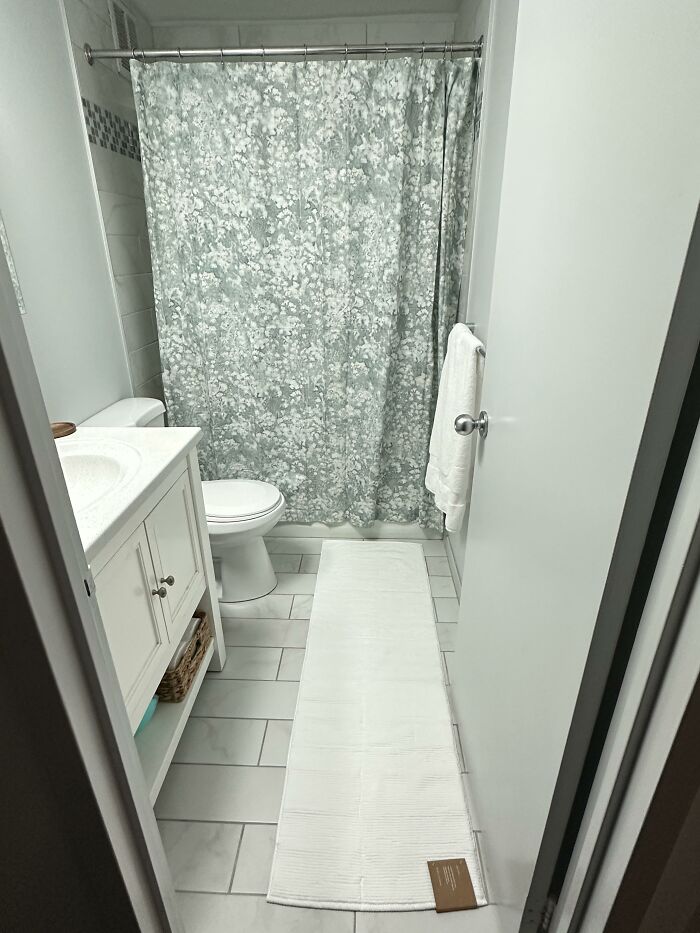 A small bathroom with a floral shower curtain, white vanity, toilet, and a long white bath mat, displaying home design choices.