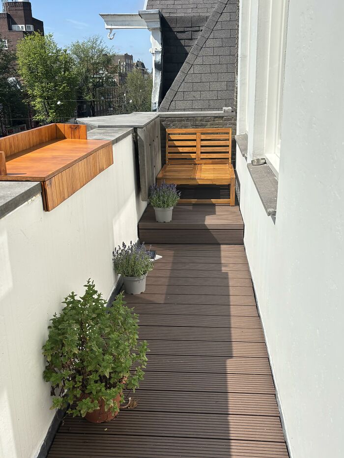A narrow outdoor rooftop terrace with dark wood flooring, a wooden bench, and potted plants, illustrating home design choices.