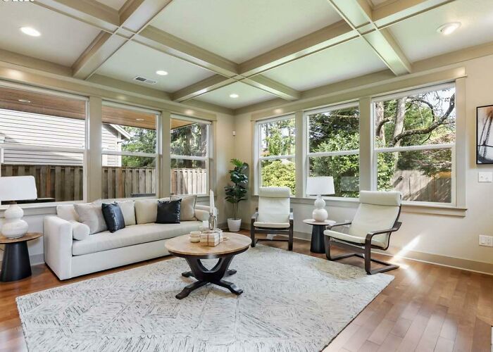 A spacious, elegant living room with a coffered ceiling, large windows, a light-colored sofa, and two armchairs, highlighting home design choices.