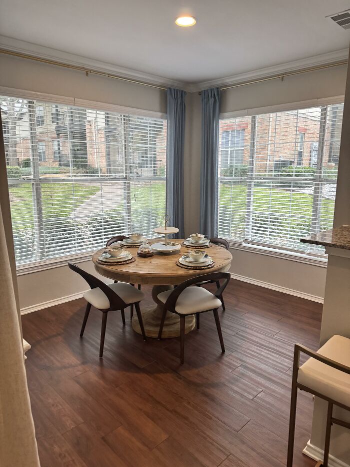 A dining nook with a round wooden table, four chairs, and large windows with blinds, displaying home design choices.