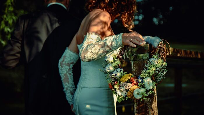 A bride with her back to the camera, wearing a white lace dress, leaning on a wooden fence with a floral wreath, capturing a wedding moment.