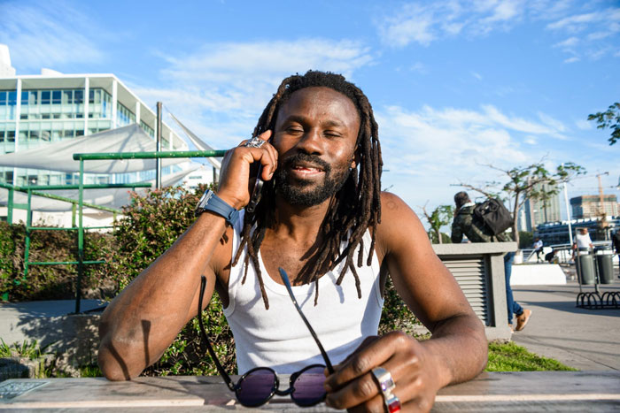 A man with dreadlocks, possibly the very large man, smiling while talking on a phone, with a bright sky in the background. A man with dreadlocks, possibly the very large man, smiling while talking on a phone, with a bright sky in the background.
