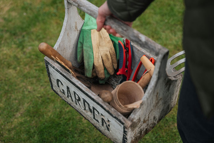 A person carries a wooden 'GARDEN' toolbox filled with gardening gloves and tools, reflecting the family free table tradition.