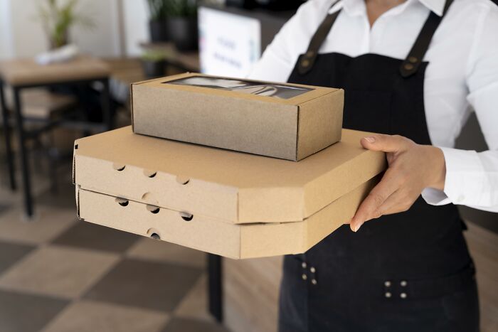 A person in an apron holding two cardboard takeaway boxes, one large and one smaller on top. These boxes represent unhinged house rules around family meals.