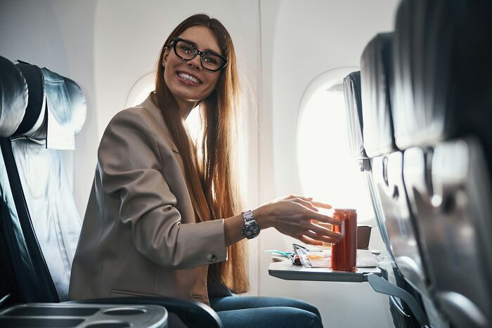 A smiling passenger on an airplane with a drink, potentially ready to ask cabin crew unhinged questions.