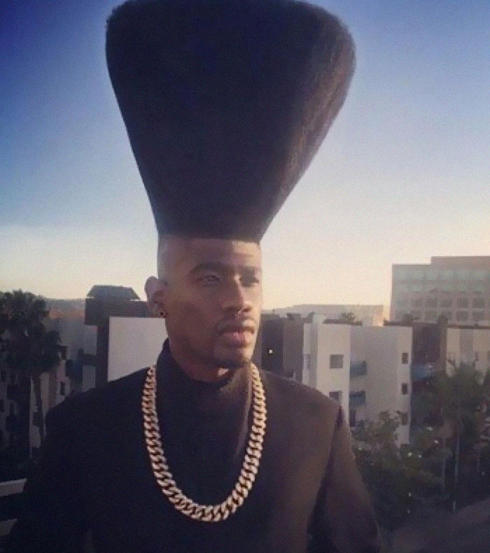 A man wearing a gold chain and a strikingly bold, tall, conical hairstyle against a city skyline.