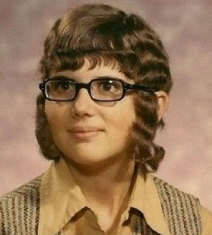 A woman with a bold, retro-inspired short hairstyle with bangs and curly sideburns, wearing glasses.