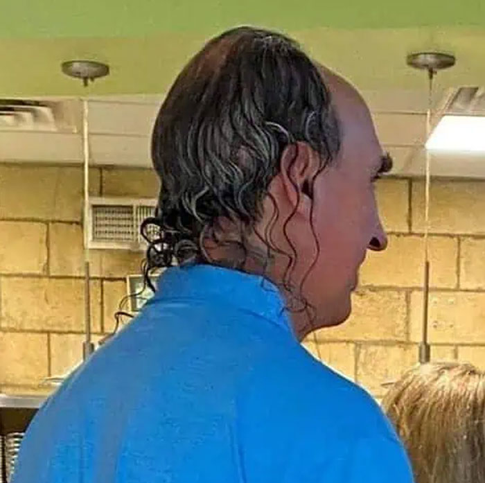 A side profile of a man with an unusual bold hairstyle, showing long, curly strands trailing from a balding head.