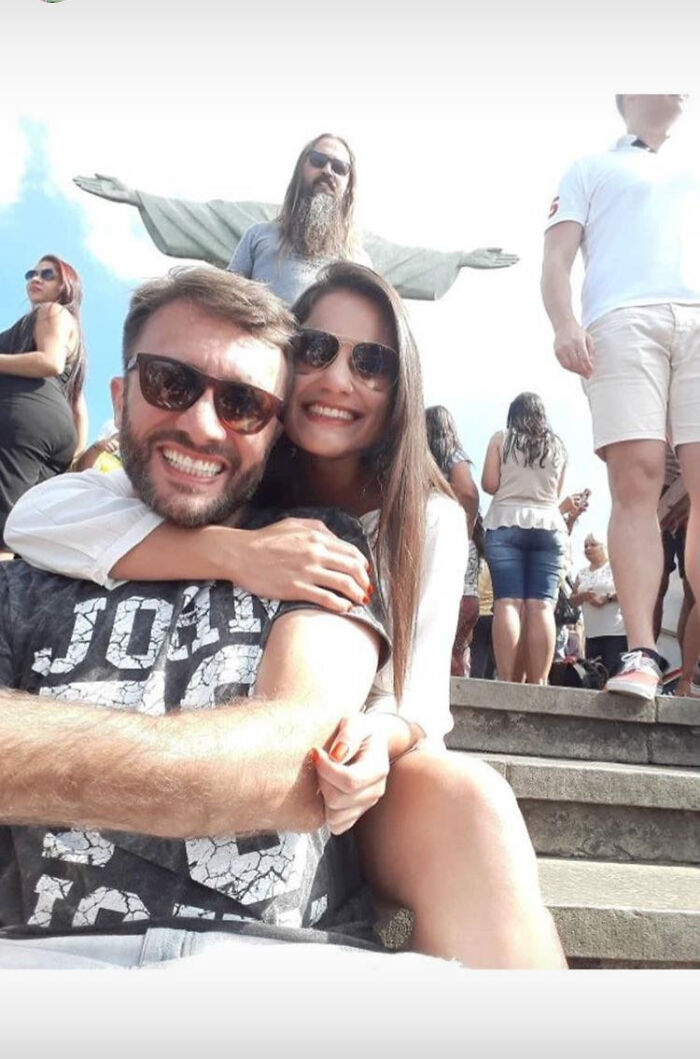 A couple smiling for a photo with a man photobombing behind them, mimicking a statue, an unexpected trip moment.