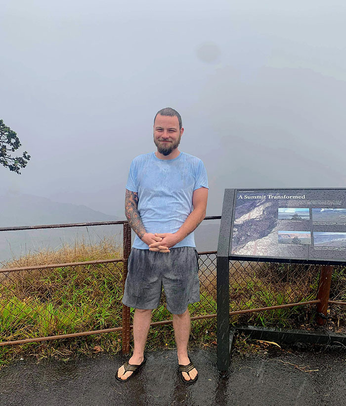 A man standing next to a sign in the rain on a mountain, depicting trips that didn't go as planned.
