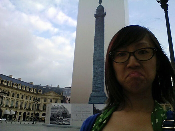 A woman with glasses and a disgruntled face taking a selfie in front of a monument, a trip surprise.