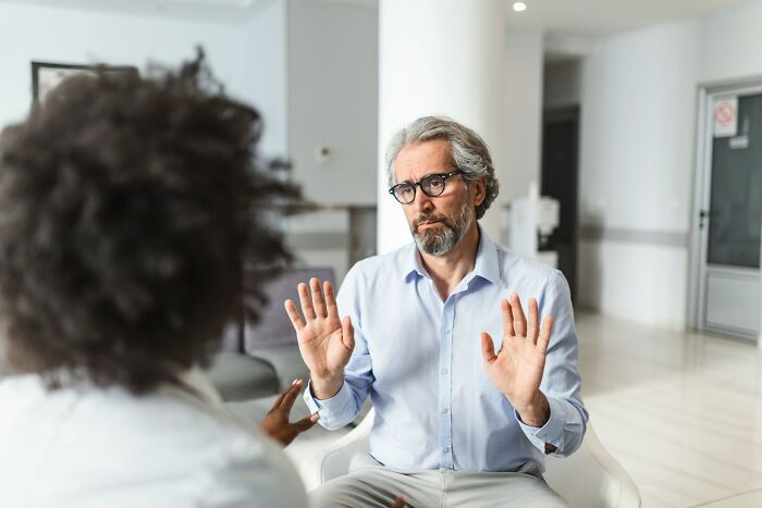 A patient with gray hair and glasses gestures with open palms while talking to a doctor about hard diagnoses.